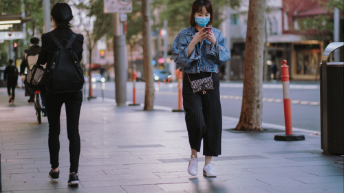 A person wearing a surgical mask looks at their phone while walking on the sidewalk