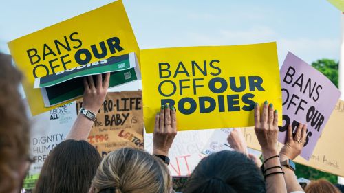 Hands hold yellow signs saying "bans off our bodies" in front of a blue sky