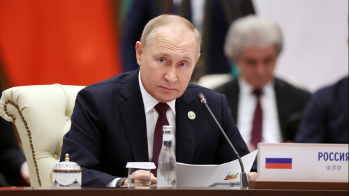 Vladimir Putin sits at a desk and looks up from a folio with raised eyebrows, red flag behind him. 