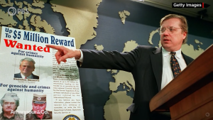 David Scheffer points to a poster with "Up to $5 million reward, wanted" in front of a blue and beige map.