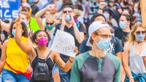 People wearing face masks march during protests; a sign in the foreground reads, 'No Justice No Peace'