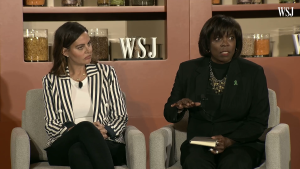 Ertharin Cousin (seated right) and Ashley Tyrner (seated left) in beige chairs in front of a wood paneled shelf wall.